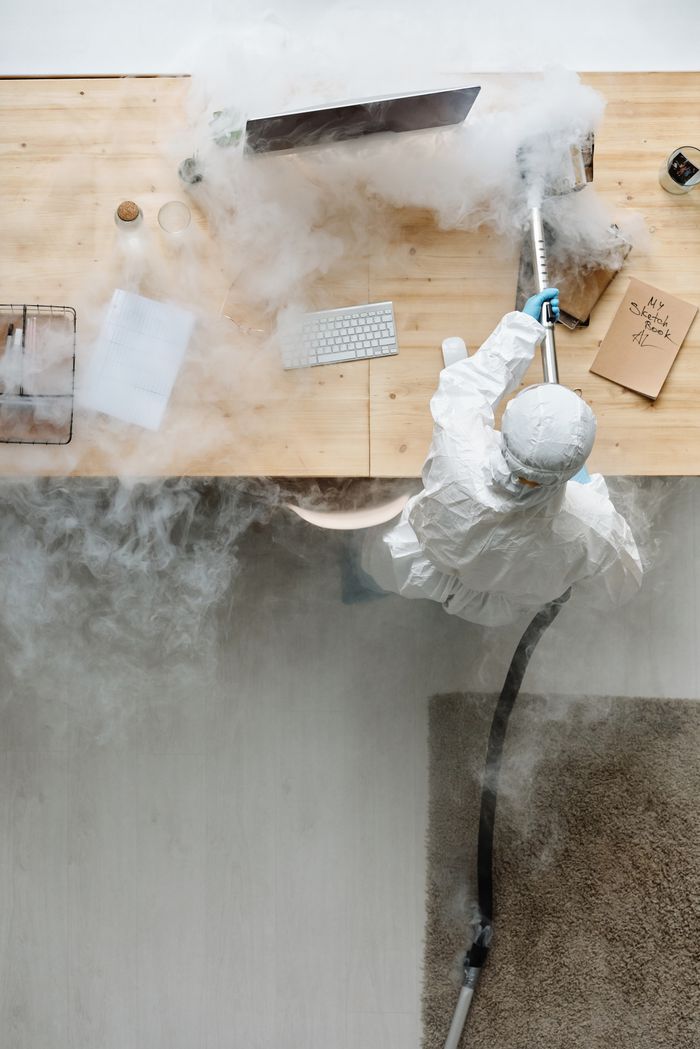 services-02 A person in protective gear sanitizing an office desk using fumigation equipment during daytime.