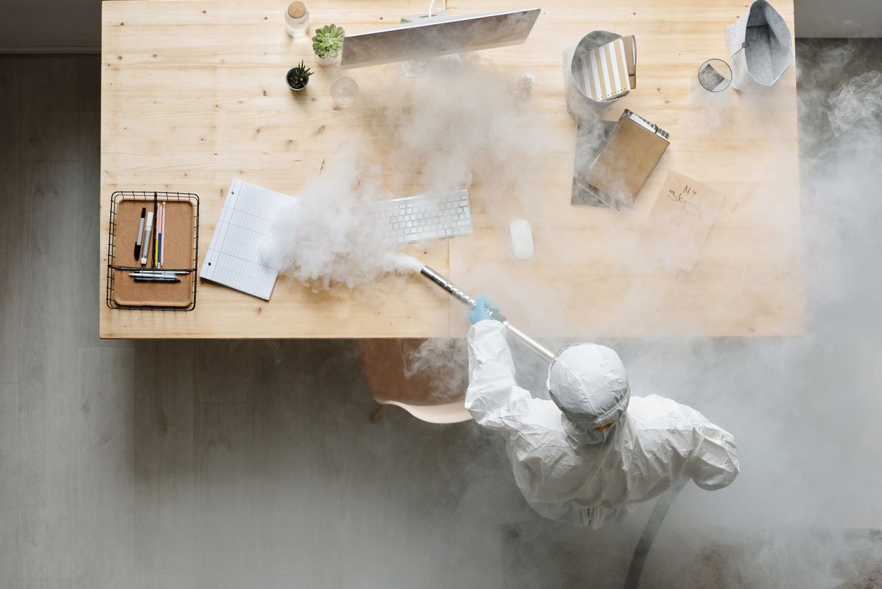 gallery-1 An individual disinfects a workspace using fumigation, wearing full protective gear for safety.