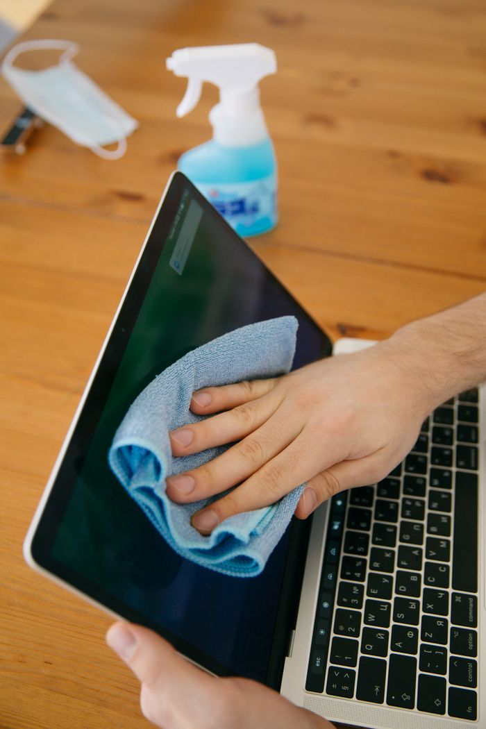 about-01 A person wipes a laptop screen using a blue cloth and spray bottle on a wooden table for hygiene.